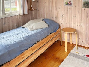 A single wooden bed with a blue mattress and pillows, a small wooden stool, and a wooden wall with a window and a picture frame. - 6 Person Holiday Home in Laholm (Skummesloevsstrand)