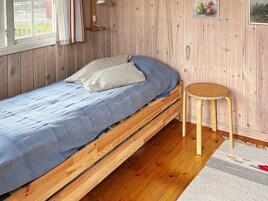 A single wooden bed with a blue mattress and pillows, a small wooden stool, and a wooden wall with a window and a picture frame.
