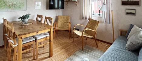 A living room with a wooden dining table, chairs, a sofa, a television, and a window with curtains.