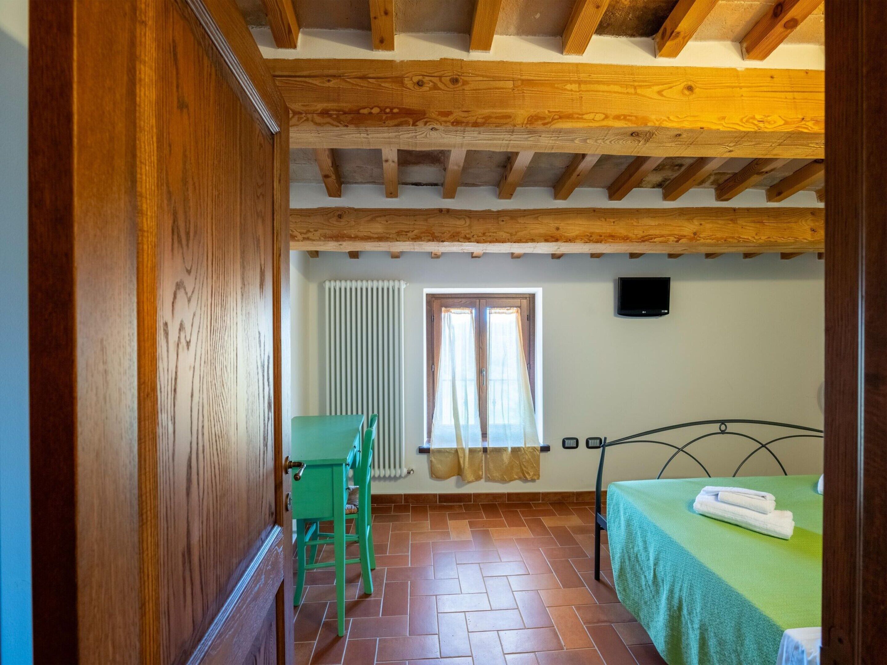 A room with a wooden ceiling, a bed with a green bedspread, a chair, a radiator, and a window with curtains.