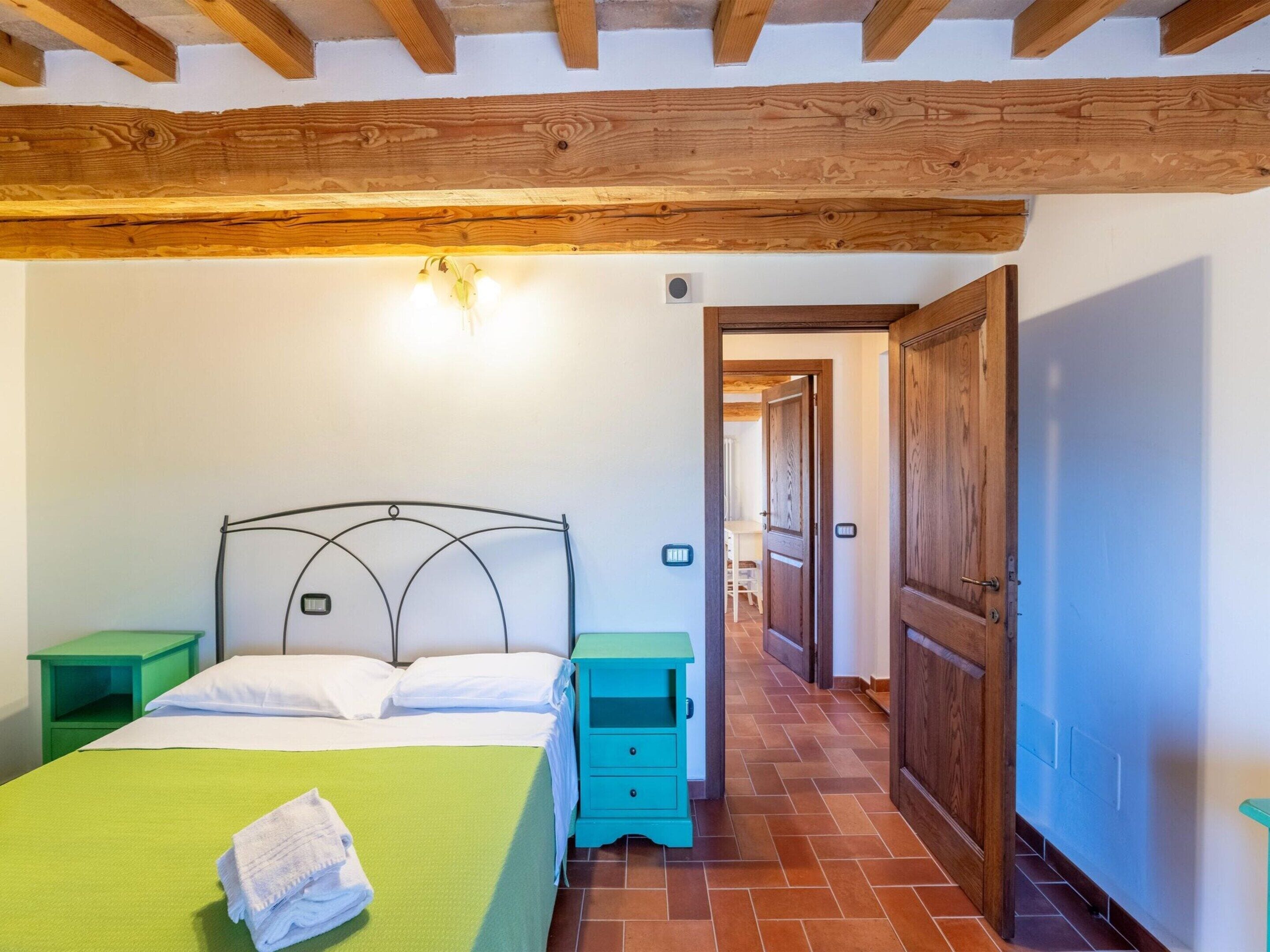 A bedroom with a bed, green nightstands, a wooden ceiling, and a door leading to another room.