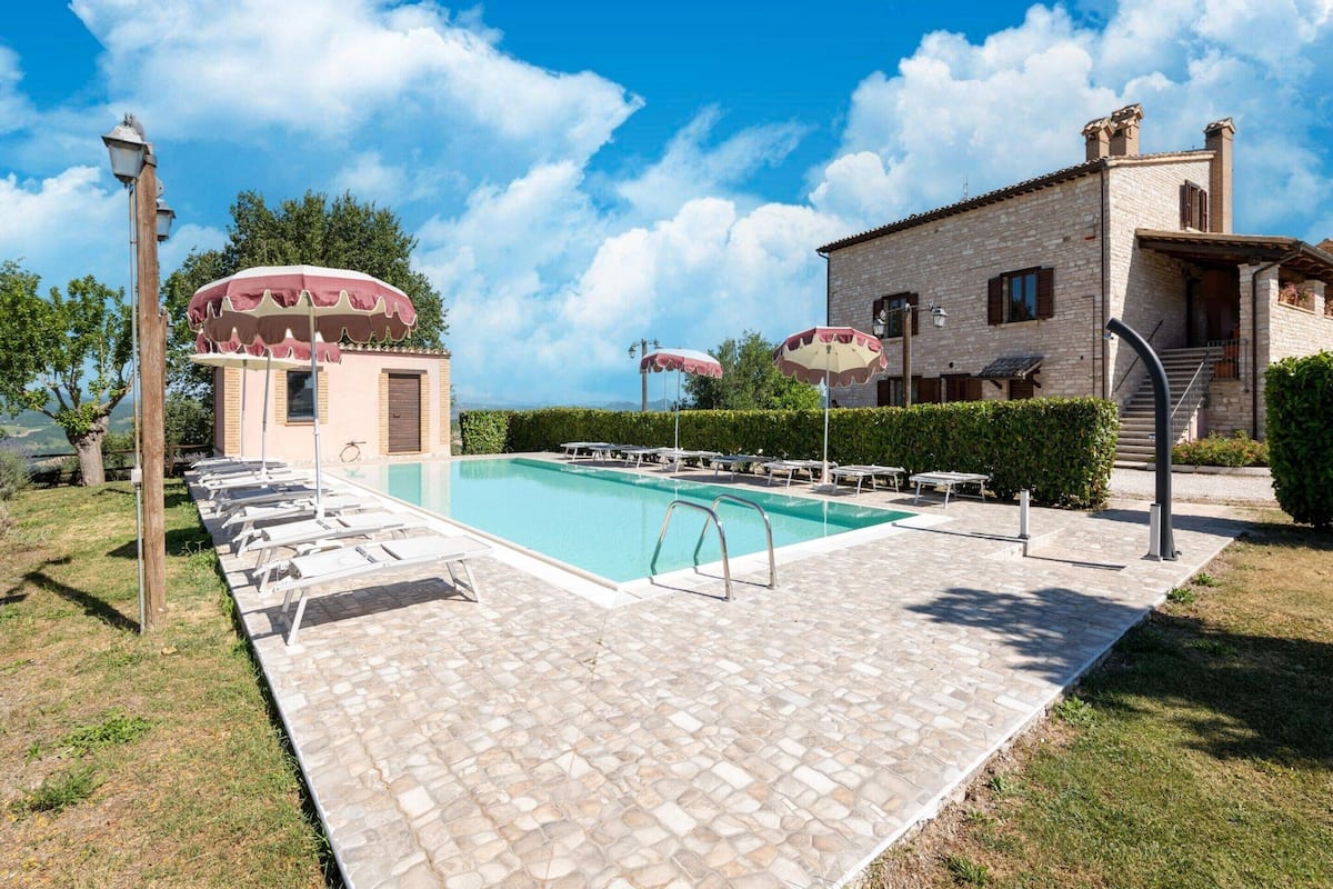 A pool area with lounge chairs and umbrellas, a stone building with a staircase, and a clear sky.