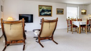 Una habitación con dos sillas de madera con asientos acolchados, una mesa de comedor con sillas, un televisor y obras de arte enmarcadas en la pared.