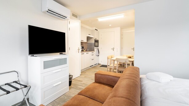 A modern hotel room with a flat-screen TV, a sofa, a dining area, and a kitchenette. - Serene Stay With All You Need (Landgraaf)