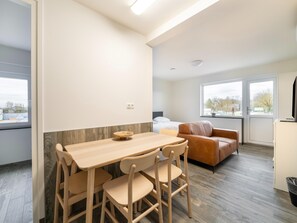A modern, well-lit room with a wooden dining table and chairs, a sofa, and large windows offering a view of the outdoors. - Serene Stay With All You Need (Landgraaf)
