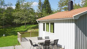A wooden deck with a table and chairs, a white building with a red roof, and a green landscape in the background. - 5 Person Holiday Home in Stromstad (Stromstad)