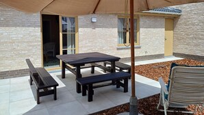 Un patio al aire libre con una mesa y bancos bajo un gran paraguas, rodeado por un muro de ladrillo y una terraza de madera.