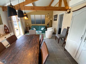 A dining area with a wooden table and chairs, a green sofa, and a white armchair. There are pendant lights above the table and a view of the outdoors through large windows. - Holiday Home in Koksijde Near Sint-andre Beach (Koksijde)