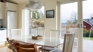 Un comedor moderno con una mesa y sillas blancas, un lámpara colgante, y vistas a un muelle y árboles a través de grandes puertas de vidrio.