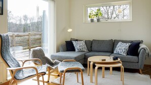 Una sala de estar acogedora con un sofá seccional gris, dos sillas de madera con cojines grises, una mesa redonda pequeña de madera y un ventanal con vistas a árboles y un edificio.