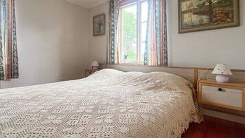 A bedroom with a large bed, a nightstand, a lamp, a window with curtains, and a painting on the wall.