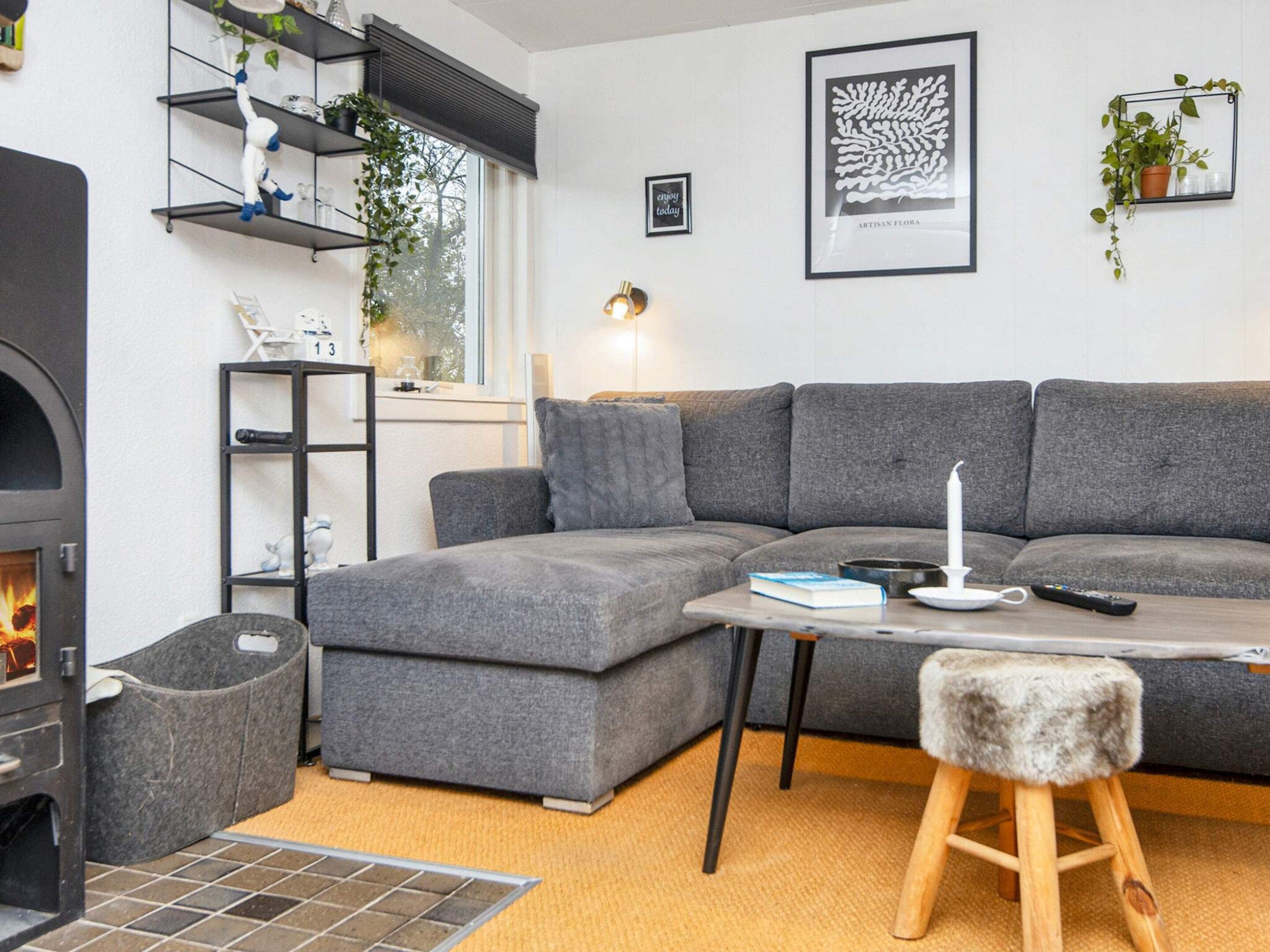 Ein modernes Wohnzimmer mit einem grauen Ecksofa, einem kleinen Tisch mit Kerze, einem Kamin und einem an der Wand befestigten Regal mit Pflanzen.