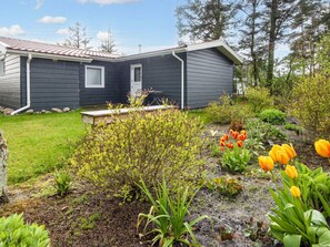 Una casa moderna con fachada oscura, un pequeño porche y un jardín con diversas plantas y flores.