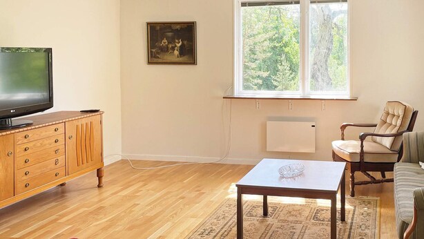 A living room with a wooden cabinet, a television, a sofa, a chair, and a large window with a view of trees. - 4 Person Holiday Home in Asarum (Asarum)