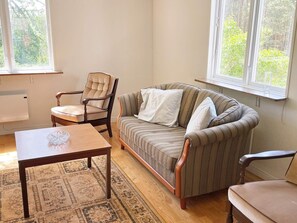 A living room with a wooden table, a striped sofa, a tufted armchair, and a view of greenery through the windows. - 4 Person Holiday Home in Asarum (Asarum)