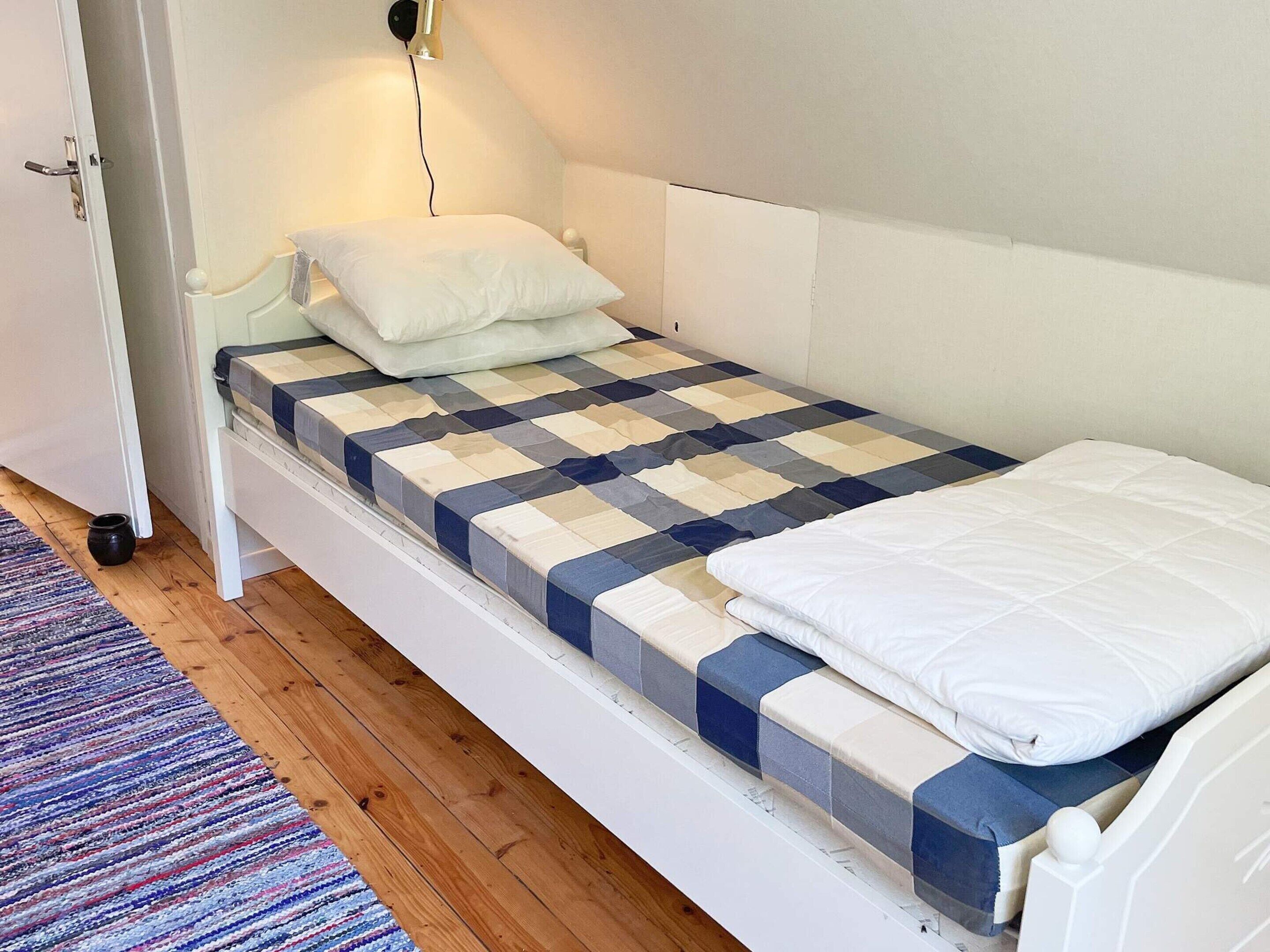 A single bed with a checkered bedspread, a white pillow, and a bedside table with a lamp.