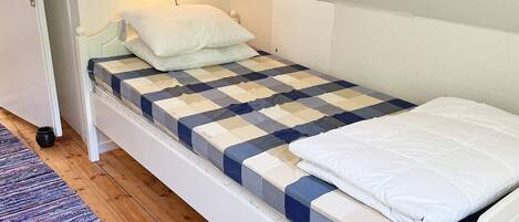 A single bed with a checkered bedspread, a white pillow, and a bedside table with a lamp.