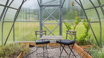 A greenhouse with a bench and chairs, a wooden table, and a view of a green field through the transparent roof.