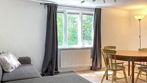 Een kamer met een bank, een eettafel met stoelen, een raam met gordijnen en een radiator.