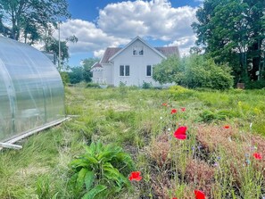 Une maison avec un jardin à l’avant, une serre sur la gauche et un ciel dégagé.