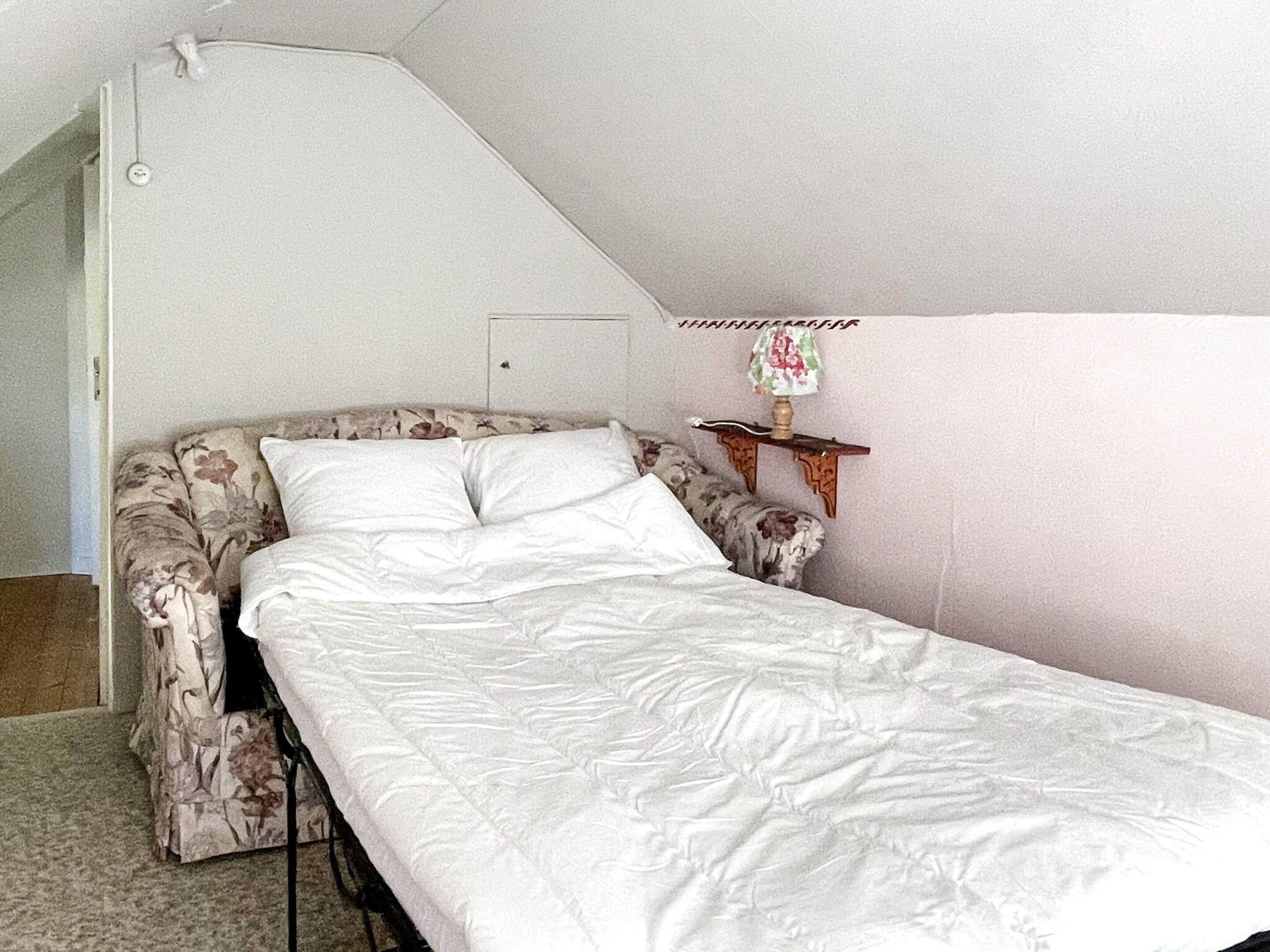 Un lit simple avec un matelas blanc et une tête de lit à motifs floraux, dans une pièce dont le plafond est en pente.