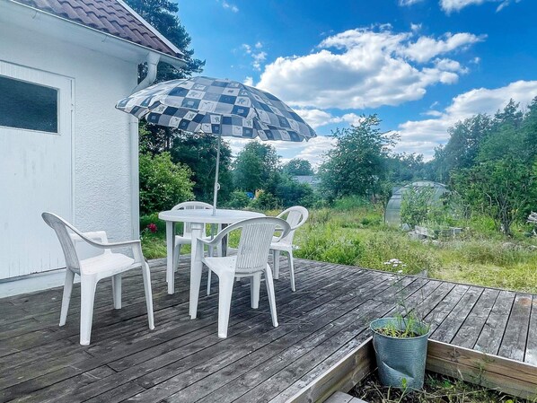 Una terraza de madera con una mesa, sillas, una planta en maceta y un edificio blanco al fondo.