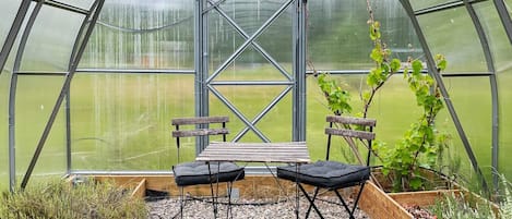 A greenhouse with a bench and chairs, a wooden table, and a view of a green field through the transparent roof.