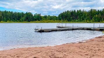 A sandy beach with a wooden dock extending into a calm lake, surrounded by dense green forest.