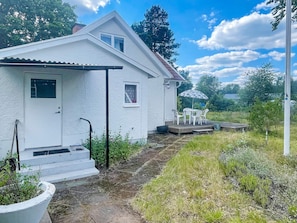 A white house with a front door, a small porch with a table and chairs, and a garden area with grass and plants. - 5 Person Holiday Home in Bockara (Bockara)