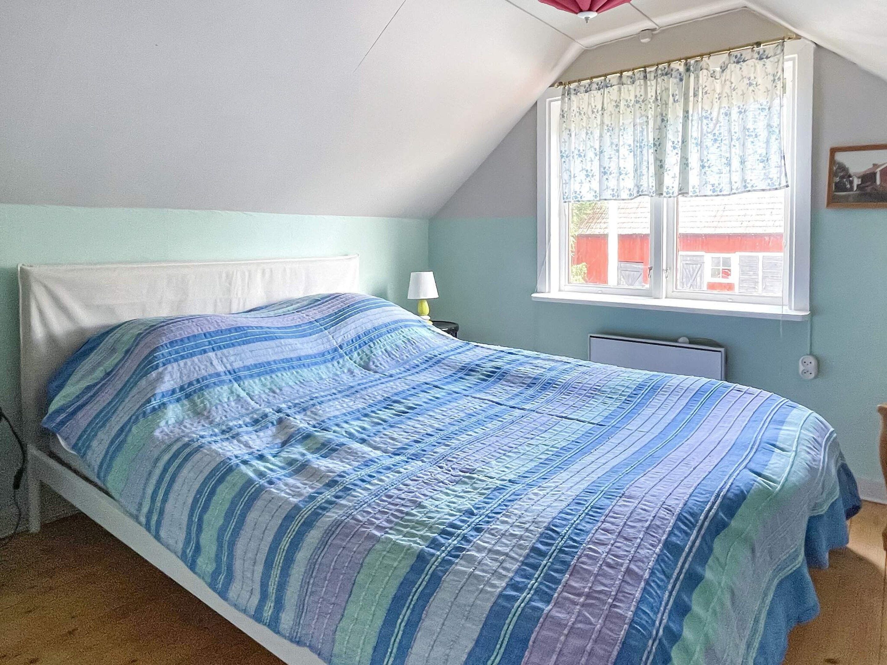 Una cama individual con una colcha a rayas en una habitación con techo inclinado y ventana con cortina.