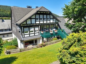 Une maison traditionnelle à colombages, dotée d’un balcon, entourée d’un jardin bien entretenu et d’une clôture en bois.