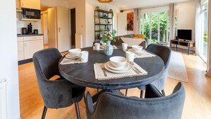 A modern dining area with a round table set for four, surrounded by dark chairs. A unique light fixture hangs above, and the room has a view of greenery through large windows. - Holiday Home in Overijssel Near Cap Fun Park (Rheezerveen)