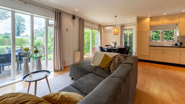 A spacious living room with a grey sofa, a small round table, and a view of the garden through large sliding doors. - Holiday Home in Overijssel Near Cap Fun Park (Rheezerveen)
