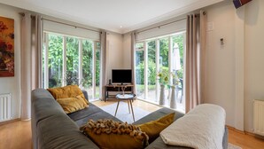 A spacious living room with a large grey sofa, a flat-screen TV on a wooden stand, and a view of a lush garden through large windows. - Holiday Home in Overijssel Near Cap Fun Park (Rheezerveen)