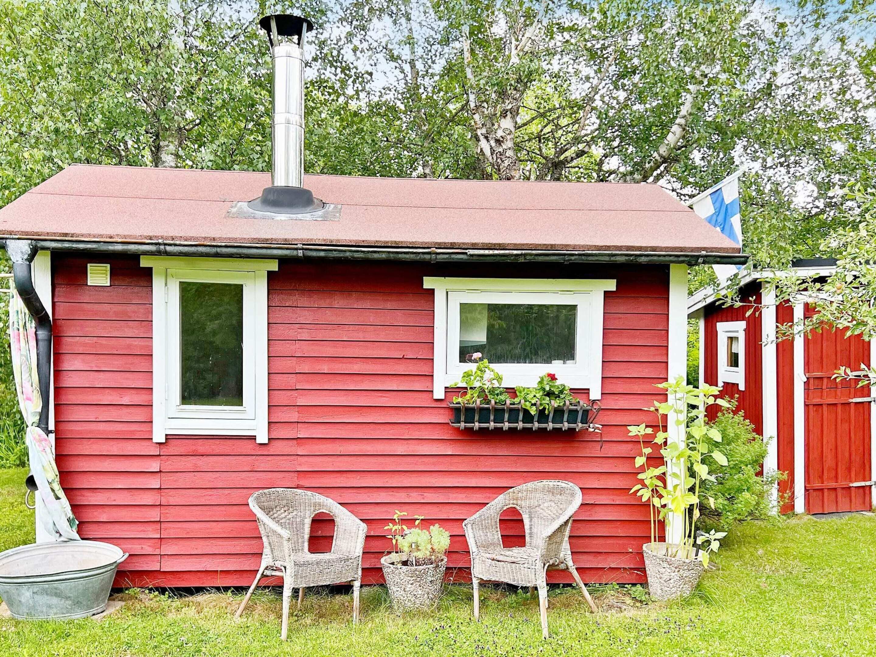 A red wooden house with two windows, a chimney, and a small porch with two wicker chairs.