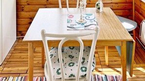 A wooden cabin interior with a dining table, white chairs, a window with a view of greenery, and a small kitchen area. - 5 Person Holiday Home in Sodertalje (Sodertalje)