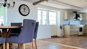 A dining area with a wooden table and chairs, a wall clock, and a kitchen with cabinets and appliances. - 8 Person Holiday Home in Skivarp (Skivarp)