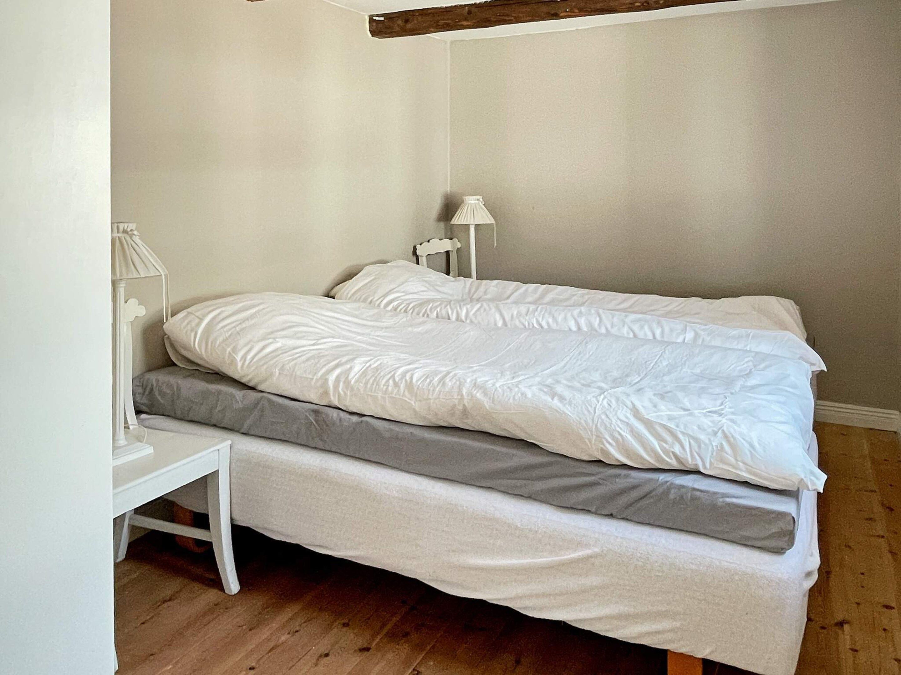 Una cama individual con colchón blanco y ropa de cama gris, una mesita de noche blanca y una lámpara blanca.