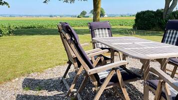 An outdoor seating area with a table and chairs, surrounded by gravel and grass, with a view of a field and a tree.
