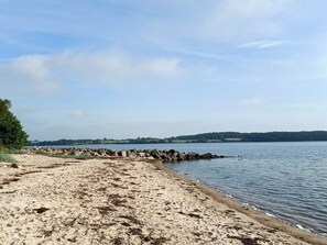 A sandy beach with a rocky breakwater, calm water, and a distant tree-covered shoreline.