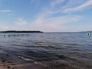 A calm body of water with a wooden dock extending into it, a distant shoreline, and a clear sky.