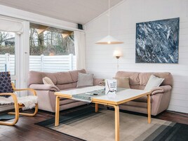 Ein gemütliches Wohnzimmer mit einem beigen Sofa, einem Holztisch und einem flauschigen Sessel. Ein Gemälde hängt an der Wand, und ein Fenster bietet Blick auf Bäume.