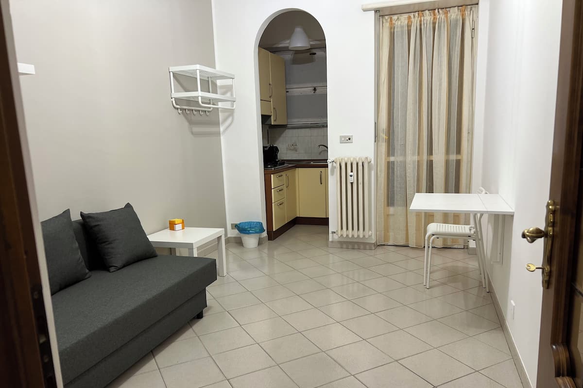A compact living space with a sofa, a small table, a radiator, a kitchen area, and a window with curtains.