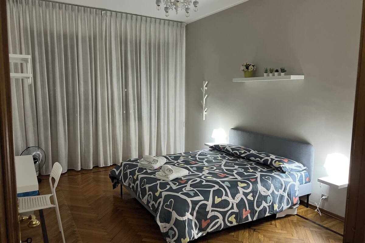 A bedroom with a bed, a chair, a desk, a chandelier, and a shelf with plants.