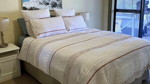 A bedroom with a large bed, white bedding, a painting of a seascape, a nightstand with a lamp, and a window with a view.