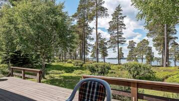 Una terraza con una silla y una mesa, con vista a un lago rodeado de altos árboles y un cielo despejado.