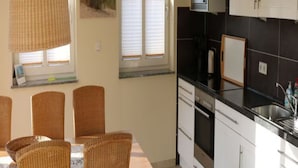A kitchen with a black countertop, white cabinets, and a built-in oven. There is a dining table with wicker chairs and a window with blinds. - Borkum Ideal 71 A Comfortable Holiday Residence (Borkum)