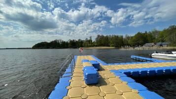 A boat dock with blue and beige floating steps leading to the water, a lifebuoy, and a distant shoreline with trees and buildings.