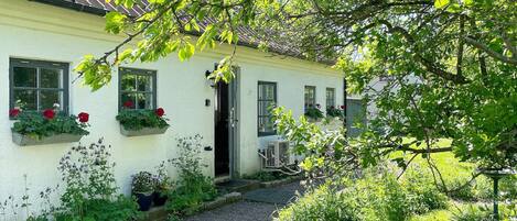 Une charmante petite maison blanche, avec un chemin de gravier menant à l’entrée, entourée d’une végétation luxuriante et de plantes en fleurs.