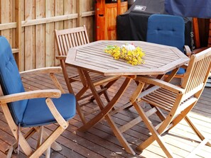 A wooden deck with a round table and four chairs, one of which has a blue cushion.